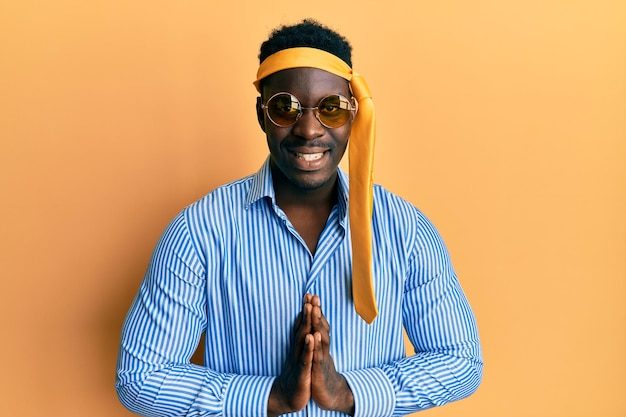 Photo gratuite bel homme noir ivre portant une cravate sur la tête et des lunettes de soleil priant avec les mains ensemble demandant pardon souriant confiant.