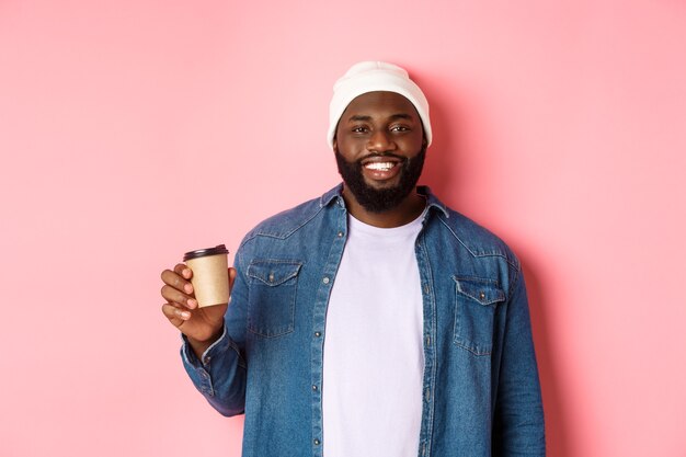 Bel homme noir moderne buvant du café à emporter, souriant et ayant l'air satisfait devant la caméra, debout sur fond rose