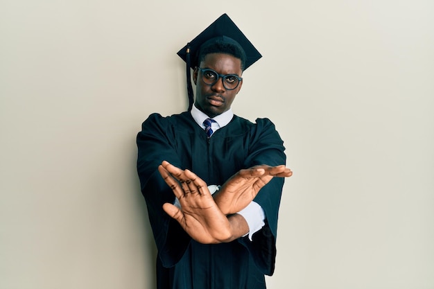 Photo gratuite bel homme noir portant une casquette de graduation et une robe de cérémonie expression de rejet croisant les bras et les paumes faisant signe négatif, visage en colère