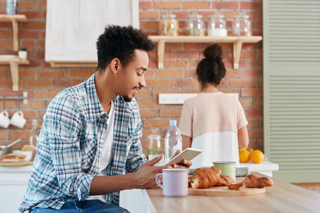 Bel homme passe la matinée à la cuisine, prend le petit déjeuner, lit un livre électronique ou des textes avec des amis à l'aide de tablette numérique