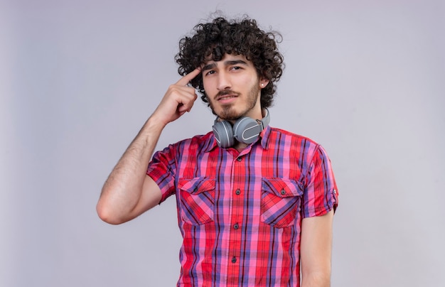 Un bel homme pensant aux cheveux bouclés en chemise à carreaux dans les écouteurs pointant sur la tête avec l'index
