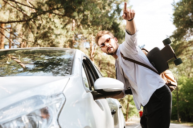 Bel homme avec planche à roulettes à l'extérieur debout près de voiture pointant.