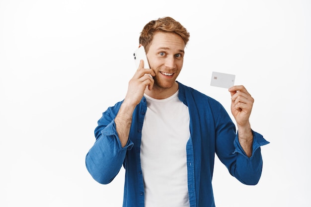 Photo gratuite bel homme rousse faisant un appel téléphonique à l'assistance bancaire, montrant une carte de crédit et souriant sans soucis, utilisant le paiement sans contact, la livraison de la commande, debout sur fond blanc