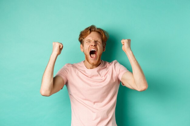 Bel homme rousse regardant un match de sport et acclamant, criant et faisant des pompes de poing, regardant l'écran et regardant la compétition, s'enracinant pour l'équipe, debout sur fond turquoise.