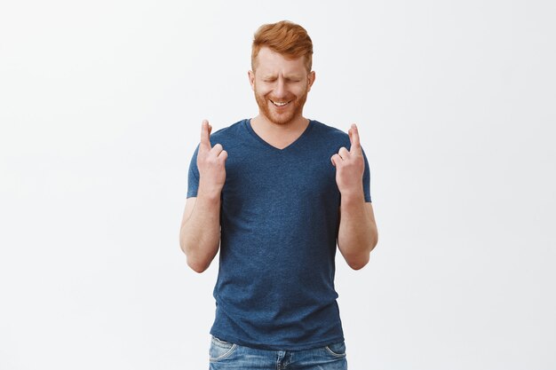 Bel homme rousse souhaitant avoir de la chance. croiser les doigts en se tenant debout sur un mur gris avec les yeux fermés et un sourire plein d'espoir, priant pour l'accomplissement du désir, portant un t-shirt bleu décontracté