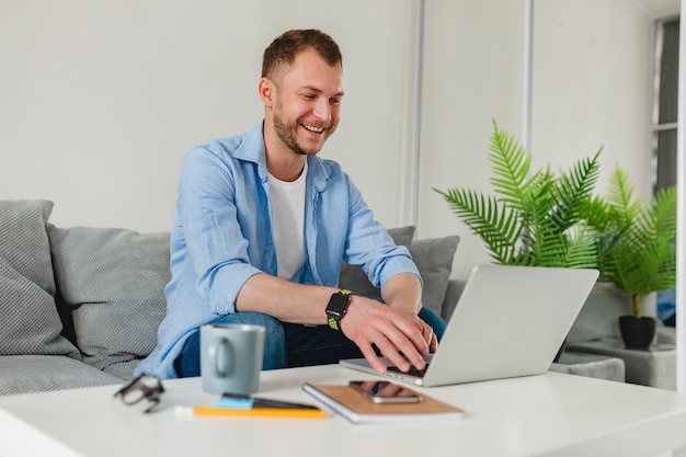 Bel Homme Souriant Assis Sur Un Canapé Buvant Du Thé à La Maison à Table Travaillant En Ligne Sur Un Ordinateur Portable Depuis La Maison