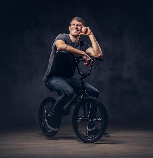 Bel homme en t-shirt et jeans assis sur un BMX. Isolé sur un fond sombre.