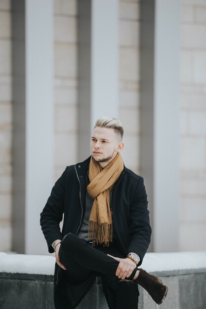 Bel homme en tenue d'hiver noire avec un foulard marron assis sur une pierre devant le bâtiment