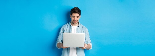 Bel homme travaillant sur un ordinateur portable souriant et regardant l'écran satisfait debout contre le backgro bleu