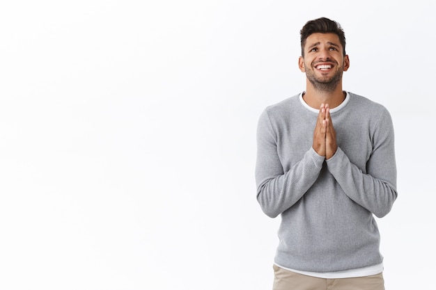 Bel homme triste et nerveux en détresse avec barbe en pull gris, serrant la main en supplication, mur blanc