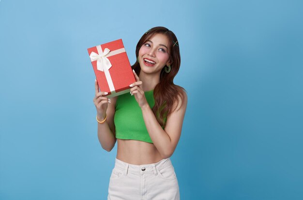 Une belle adolescente asiatique heureuse sourit avec une boîte à cadeaux rouge isolée sur un fond bleu