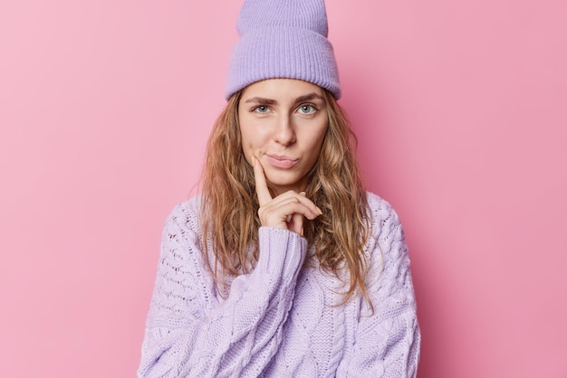 Photo gratuite belle adolescente aux cheveux longs réfléchit à quelque chose qui garde le doigt sur les joues étant profondément dans ses pensées porte un pull chaud et un chapeau isolés sur fond de studio rose. laisse-moi penser