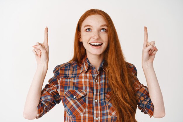 Belle adolescente aux cheveux longs rouges, regardant et pointant les doigts émerveillés, debout sur un mur blanc