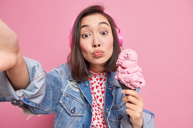 Une belle adolescente romantique d'apparence orientale tient un gros cône de glace étend le bras pour faire un selfie porte un casque écoute de la musique vêtue d'une veste en jean isolée sur un mur rose