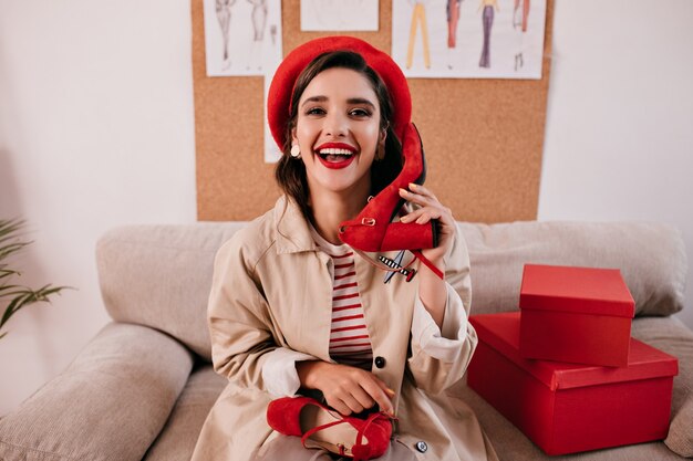 Belle belle dame en béret à la mode brillant et trench d'automne beige s'amusant dans son appartement et détient des chaussures rouges.