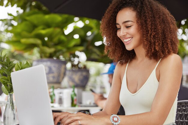 Belle belle femme heureuse aux cheveux noirs bouclés, habillée avec désinvolture, claviers sur ordinateur portable, travaille en freelance dans une cafétéria en plein air