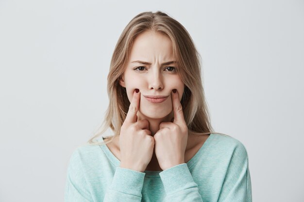 belle belle jeune femme aux cheveux blonds teints, garde les mains sur les joues, a une expression réfléchie et sérieuse, insatisfaite et mécontente