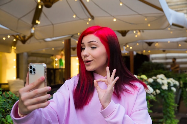 Belle blogueuse influenceuse aux cheveux roux au café parlant en faisant un appel vidéo à l'aide d'un smartphone avec ses abonnés sur les réseaux sociaux.