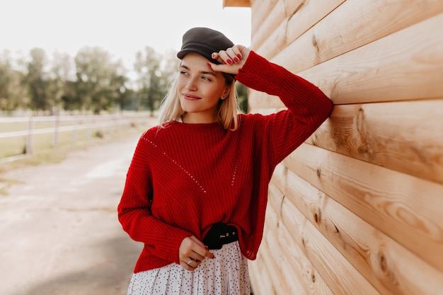 Belle blonde souriante joyeusement près de la maison en bois dans le parc. Belle fille posant dans les beaux vêtements de saison à l'extérieur.