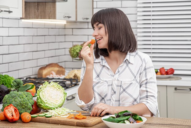 Belle brune sourit et coupe les légumes sur une salade sur le fond d'un intérieur de cuisine moderne.