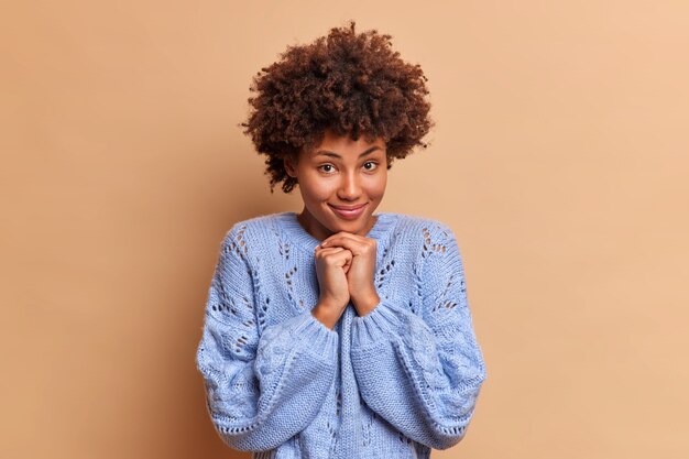 Belle charmante jeune femme aux cheveux bouclés garde les mains sous le menton a des sourires tendres et regarde directement à l'avant porte des poses de chandail décontracté contre le mur marron