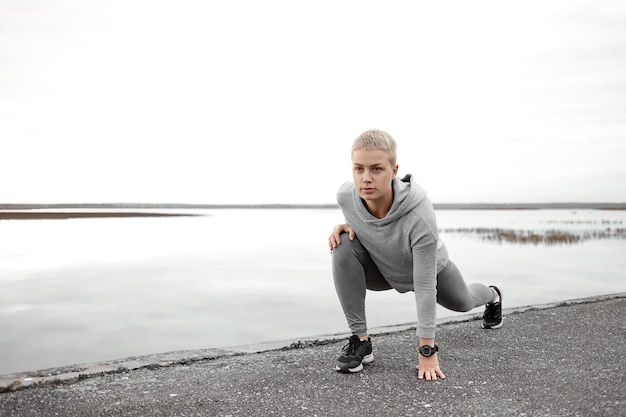 Belle coureuse caucasienne autodéterminée en vêtements de sport à la mode posant isolée sur la rive du fleuve, étirement des jambes, échauffement des muscles avant de courir à l'extérieur tôt le matin