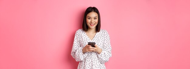 Belle dame asiatique lisant un message et souriant à l'aide d'un téléphone portable debout dans une jolie robe contre p