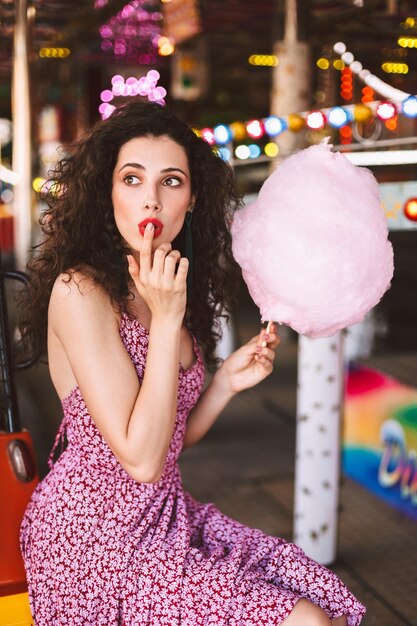 Belle dame aux cheveux bouclés noirs en robe assise avec de la barbe à papa à la main et regardant rêveusement de côté tout en passant du temps dans un parc d'attractions