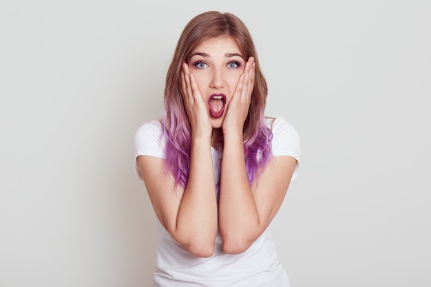 Photo gratuite belle dame aux cheveux lilas exprimant son étonnement, voit quelque chose de surprenant, entend des nouvelles choquantes, garde la bouche ouverte, tient les paumes sur les joues, posant isolée sur un mur gris.