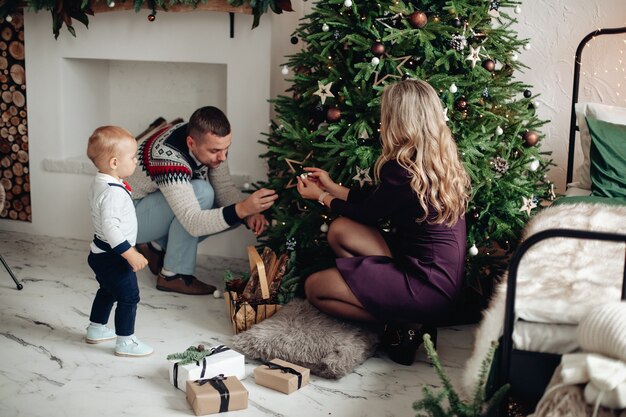 Belle dame blonde avec son mari et son enfant mignon assis près de l'arbre de Noël tout en le décorant