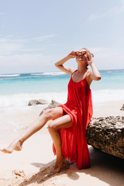 Belle dame caucasienne en longue tenue rouge se détendre à la plage. Magnifique femme bronzée se détendre près de l'océan et posant sur la pierre.