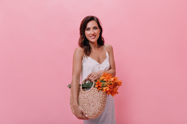 Belle dame en chemisier blanc tient un sac de paille et des fleurs orange. Jolie femme en tenue à la mode légère tient le sac à main tricoté avec bouquet.