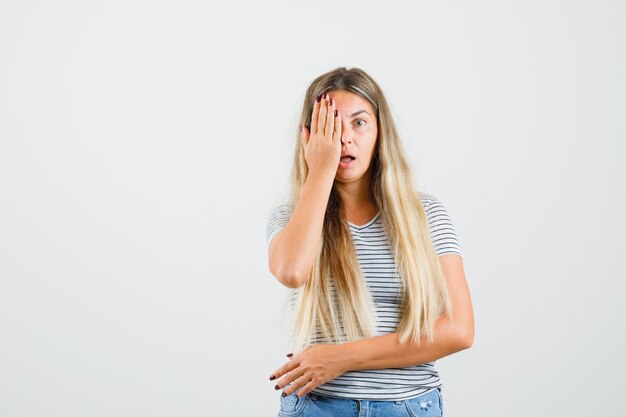 Belle dame couvrant sa moitié du visage avec la main en t-shirt et à la peur, vue de face.