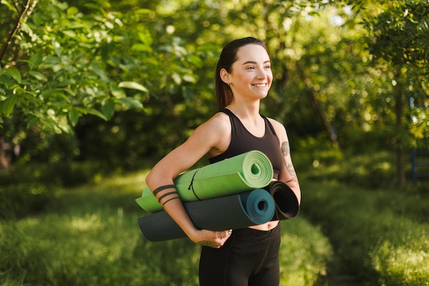 Belle dame en haut sportif noir et leggings debout avec des tapis de yoga dans les mains et regardant joyeusement de côté dans le parc