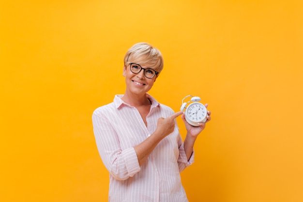 Belle dame à lunettes se penche sur la caméra et pointe vers un réveil sur fond orange