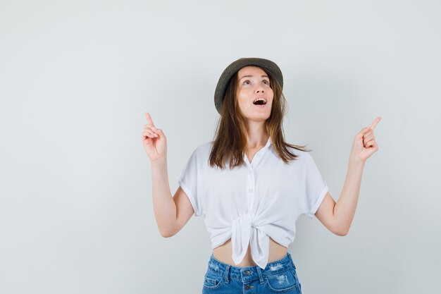 Belle dame pointant vers le haut en chemisier blanc, chapeau et regardant étonné, vue de face.