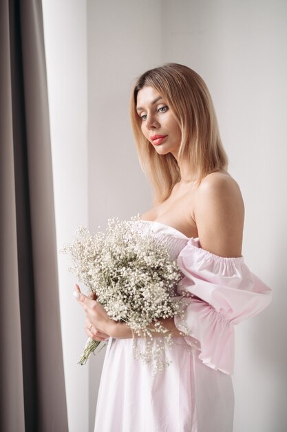 Belle dame en robe tenant un bouquet de fleurs tout en le regardant dans le salon à la maison. Notion romantique. Concept de mode de vie