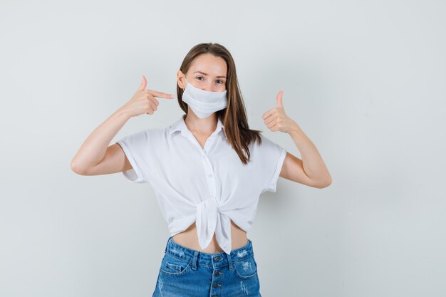 Belle dame se montrant tout en montrant le pouce vers le haut en chemisier blanc, masque facial et à la vue de face confiante.