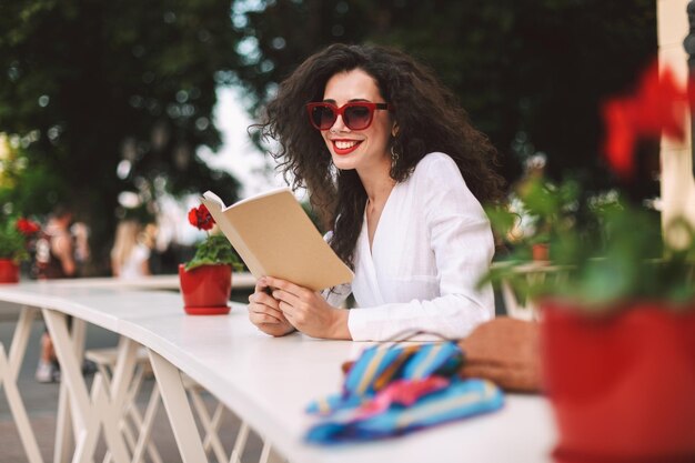 Belle dame souriante aux cheveux bouclés noirs dans des lunettes de soleil debout avec un bloc-notes dans les mains tout en passant du temps sur la terrasse d'été du café