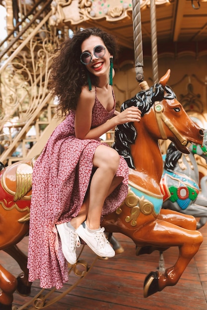 Photo gratuite belle dame souriante aux cheveux bouclés noirs en lunettes de soleil et robe assise sur un cheval de carrousel tout en passant du temps dans un parc d'attractions