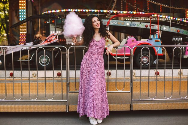 Belle dame souriante aux cheveux bouclés noirs en robe debout avec de la barbe à papa rose à la main et regardant joyeusement de côté avec une attraction de voiture sur fond dans un parc d'attractions