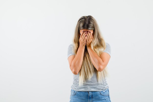 Belle dame en t-shirt couvrant son visage avec ses mains et regardant en colère, vue de face.