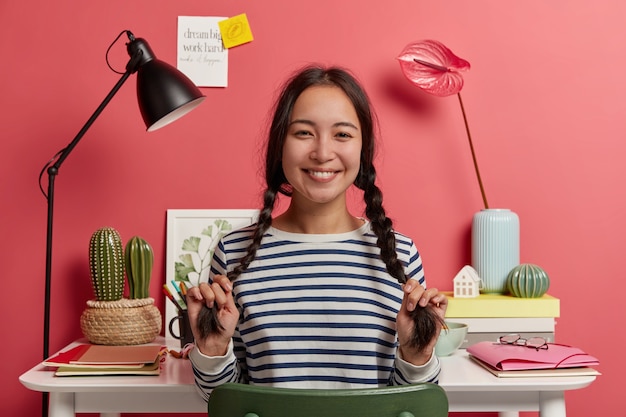 Une belle écolière s'amuse en se préparant aux examens, a deux longues tresses, sourit joyeusement, vêtue d'un pull rayé, planifie le calendrier pour le mois prochain