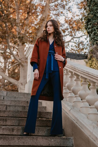 Belle élégante femme maigre souriante aux cheveux bouclés marchant dans les escaliers de la rue vêtue d'un manteau brun chaud et d'un costume bleu, style de rue à la mode automne