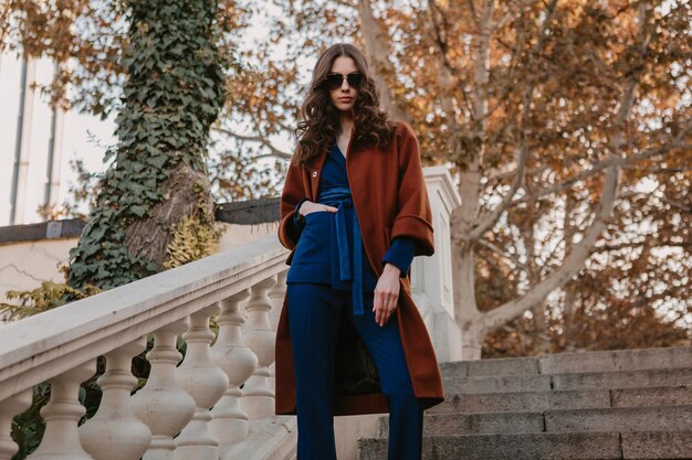 Belle élégante femme maigre souriante aux cheveux bouclés marchant dans les escaliers de la rue vêtue d'un manteau brun chaud et d'un costume bleu, style de rue à la mode automne