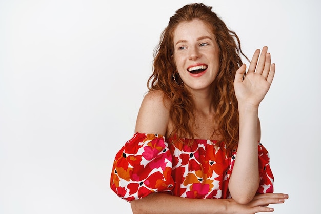 Belle et élégante femme rousse agitant la main regardant de côté et riant en disant bonjour debout en robe sur fond blanc