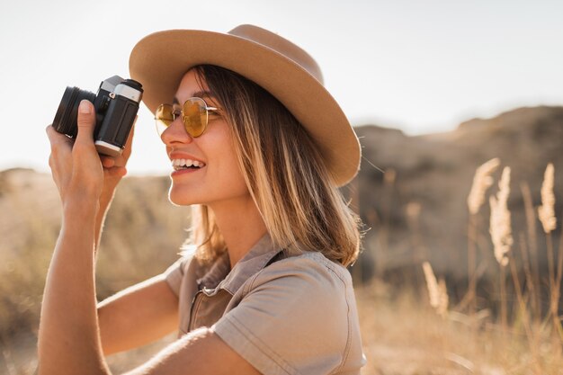 Belle élégante jeune femme en robe kaki dans le désert voyageant en Afrique en safari portant chapeau prenant photo sur appareil photo vintage