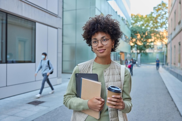 Photo gratuite une belle étudiante bouclée tient une tablette numérique pour ordinateur portable et un café à emporter concentré dans des poses à distance en milieu urbain porte des vêtements décontractés pose dans une rue animée en route vers l'université