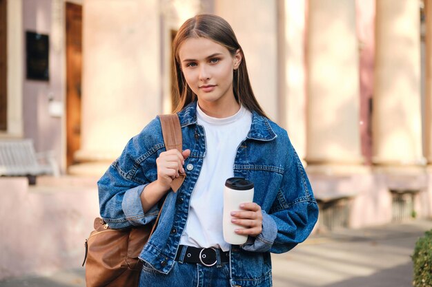 Belle étudiante décontractée élégante en veste en jean avec sac à dos regardant rêveusement à huis clos en plein air