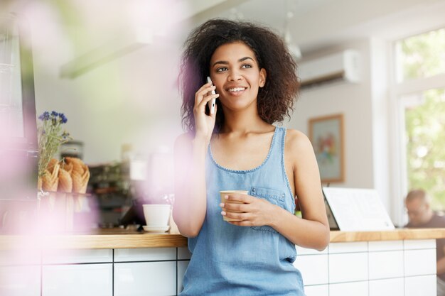 Belle étudiante gaie jeune femme africaine souriant parlant au téléphone, boire du café au café.